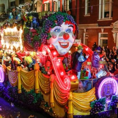 Colorful Krewe of Bacchus Mardi Gras float at night, glowing with purple, green and gold lights as costumed riders throw beads to a packed New Orleans street.