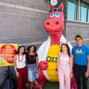 Four smiling young people pose next to Dr. Ozzie, a large red dragon statue wearing a white lab coat and stethoscope at Luskin Orthopaedic Institute for Children. A yellow sign nearby invites visitors to “Take a photo with me, Dr. Ozzie!”