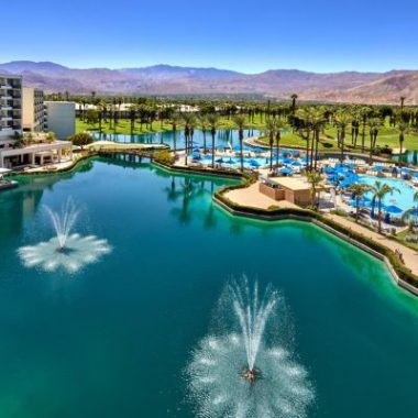 Aerial view of the JW Marriott Desert Springs Resort & Spa in Palm Desert, featuring sparkling lagoons, fountains, swimming pools, palm trees and mountain views beneath a clear blue sky.