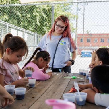 Children sit at a picnic table during a LA’s BEST summer program activity while a staff member stands nearby and supervises.