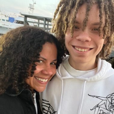 A mother with dark curly hair leans her head on her teen son's shoulder. He has blond locks. They are both smiling.