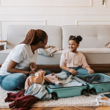 A mother and daughter pack a suitcase while sitting on a living room floor.