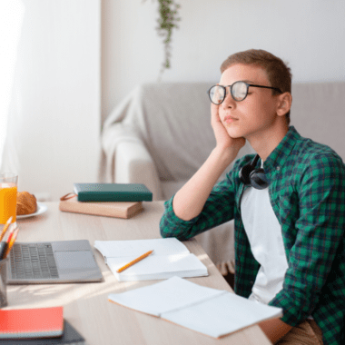 A young teen boy with closed eyes behind glasses sits at a desk spread with notebooks and papers.